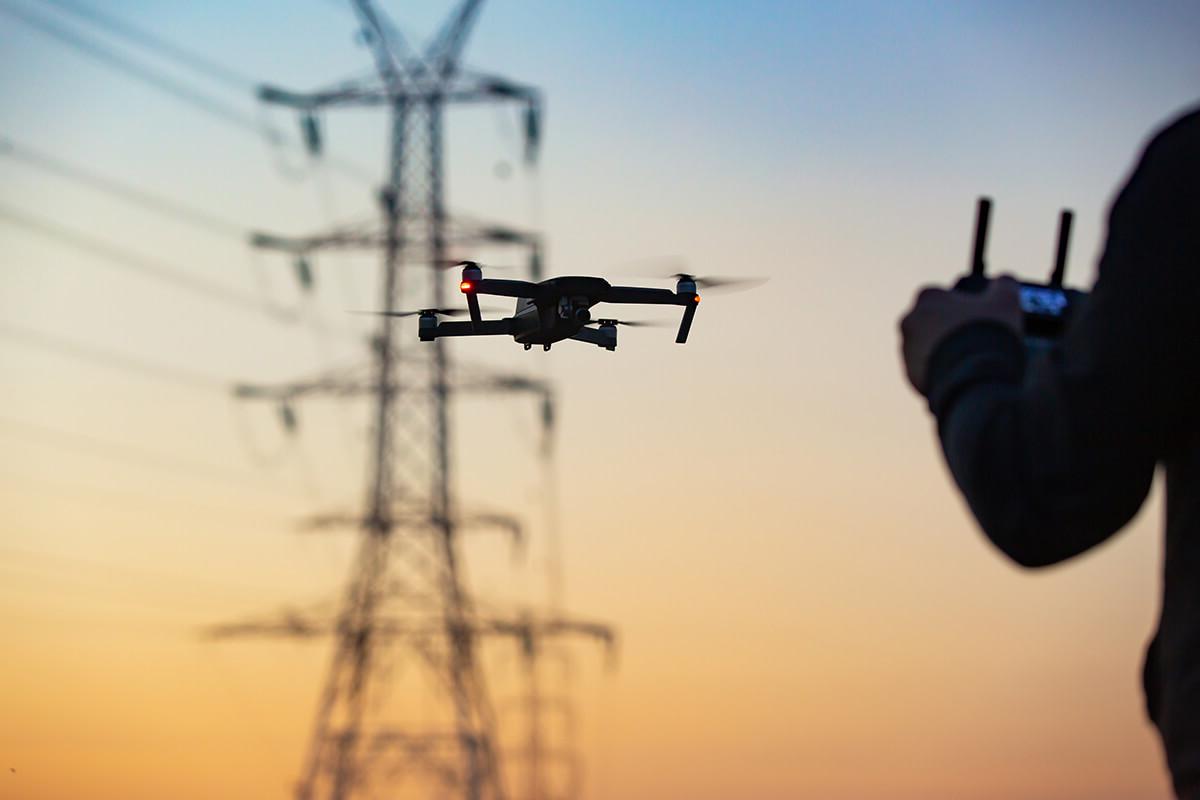 A drone flying in front of an electricity mast at sunset, with part of the pilot using the remote in the foreground